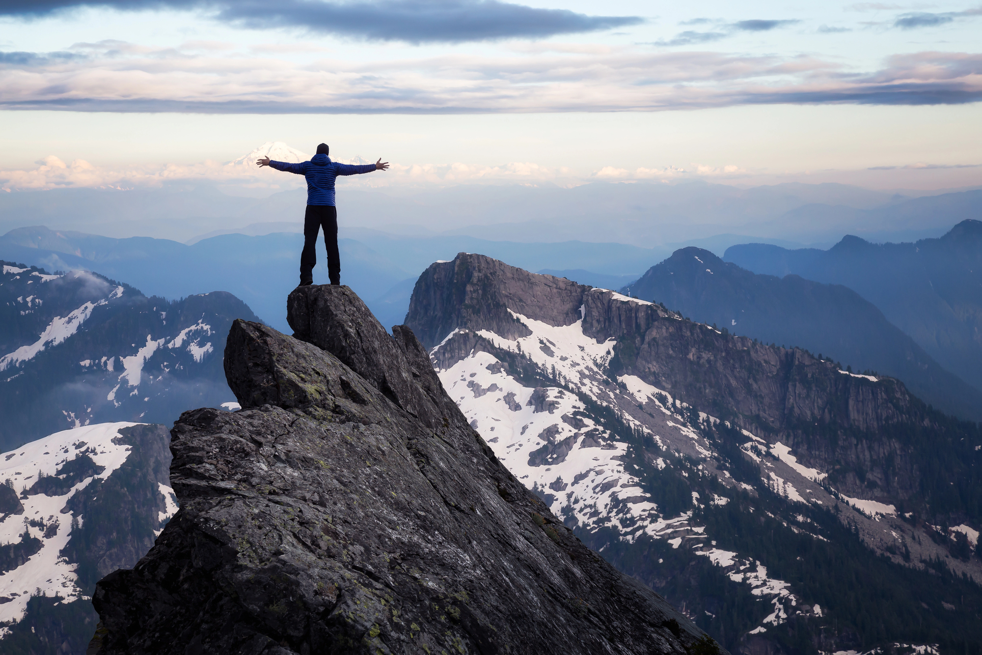 Person standing triumphantly on mountain peak overlooking snow-capped mountains